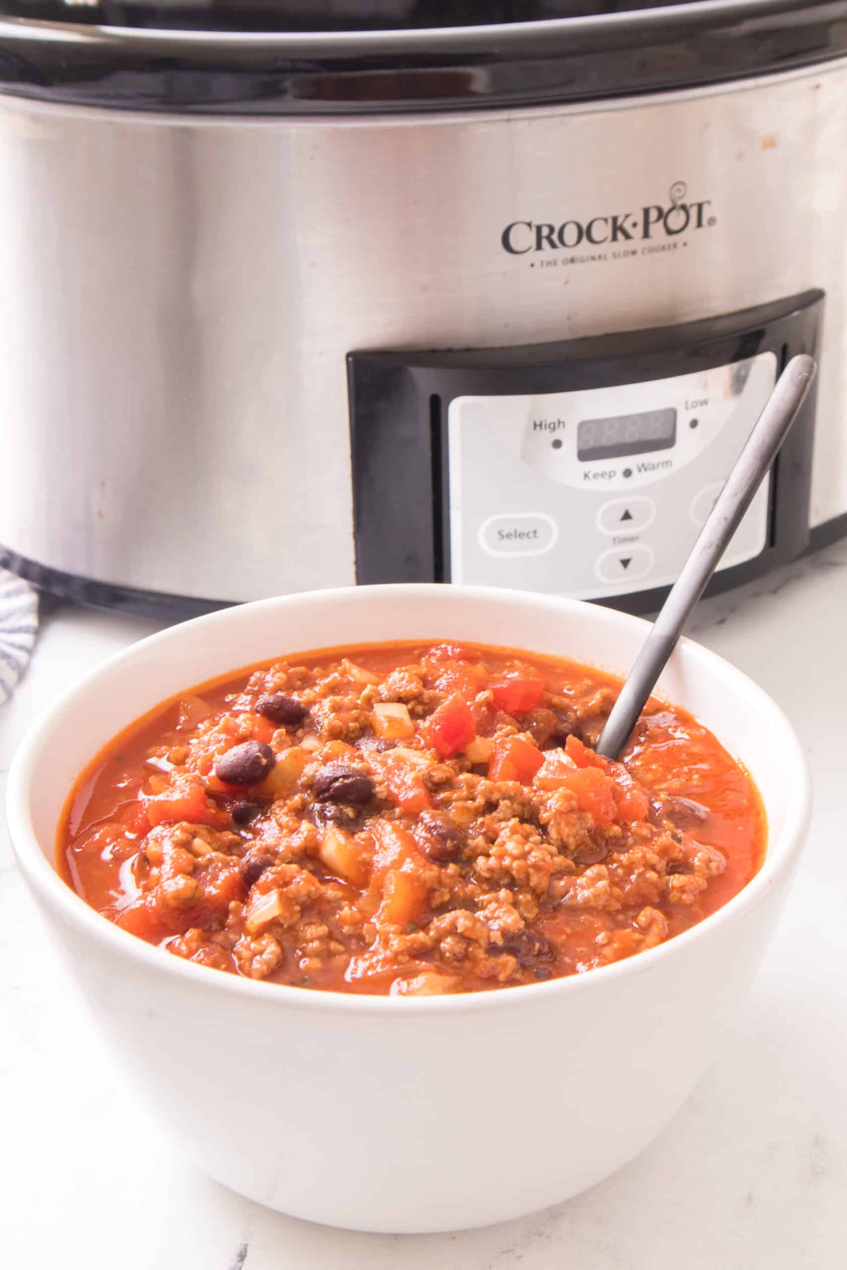 close up of chili in a white bowl with spoon and slow cooking in the background