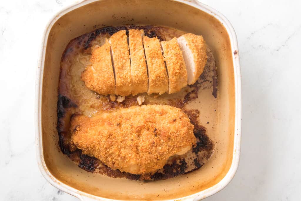 parmesan chicken cooked in baking dish with two pieces of chicken breasts, one sliced