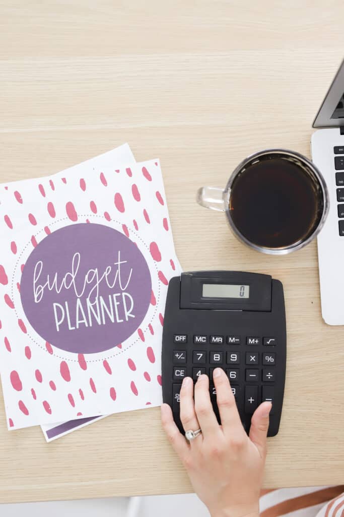 this photo shows a desktop with a printable budget planner and a hand on a calculator with a cup of coffee, showing the cover of the free budget planner offered on my blog.