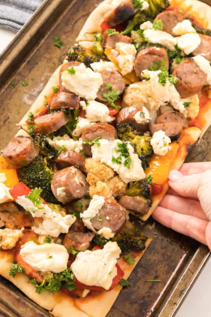 hand grabbing a slice of sausage flatbread pizza on baking sheet
