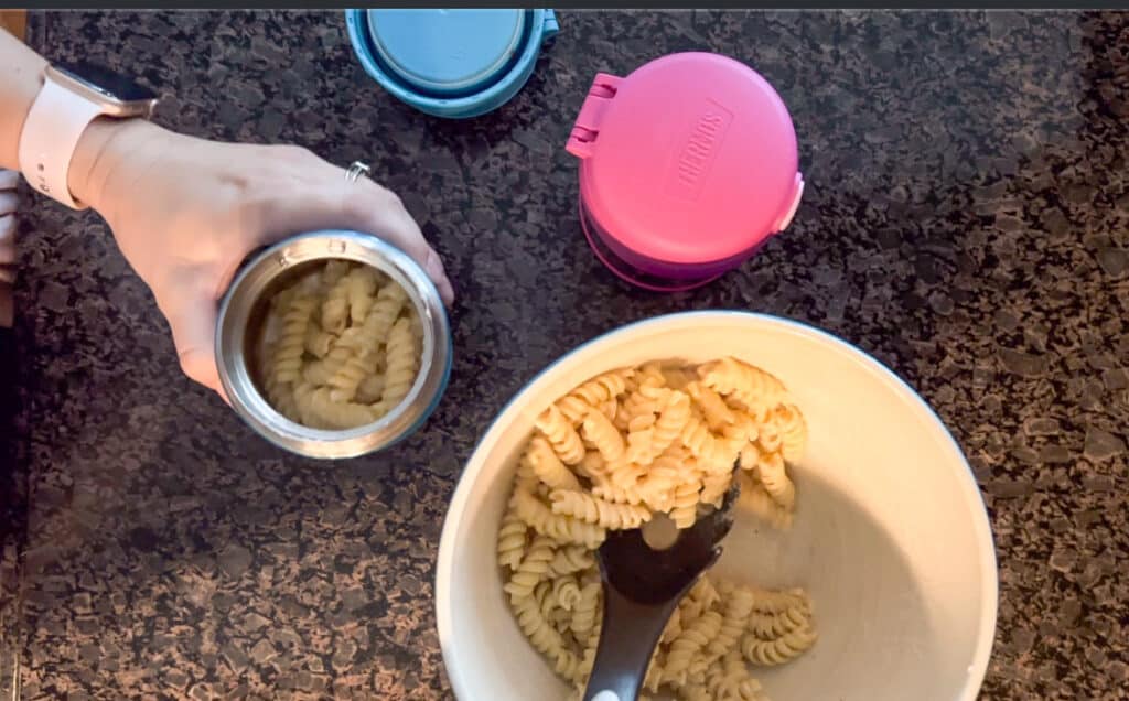 This image shows a blue and pink Thermos funtainer insulated food containers on a kitchen counter next to a bowl of premade pasta. The pasta is being spooned into the blue container. 