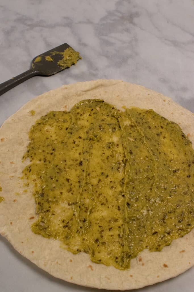 A large tortilla with pesto and mayo mixture spread in a thin layer.