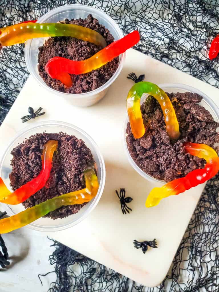 Clear plastic cups with layered chocolate pudding and Oreo cookie crumbs with gummy worms on top creating a popular kid friendly Halloween worms in dirt recipe.