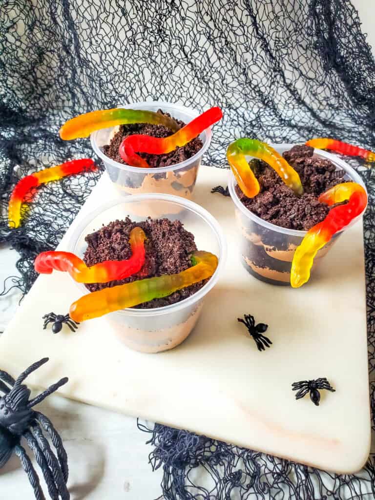 Clear plastic cups with layered chocolate pudding and Oreo cookie crumbs with gummy worms on top creating a popular kid friendly Halloween worms in dirt recipe.