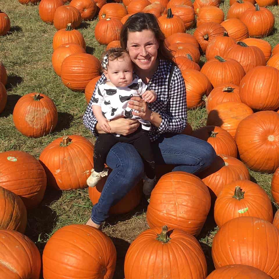 A photo of a mom and toddler girl posing with pumpkins in a pumpkin patch in fort worth. 